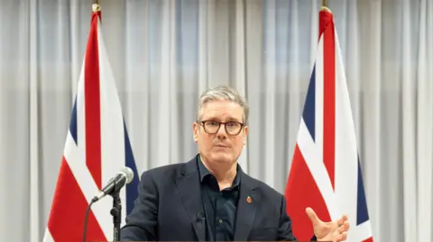 PA Media UK Prime Minister Sir Keir Starmer in dark shirt and dark suits stands in frnt of two Union Jack flags to give speech