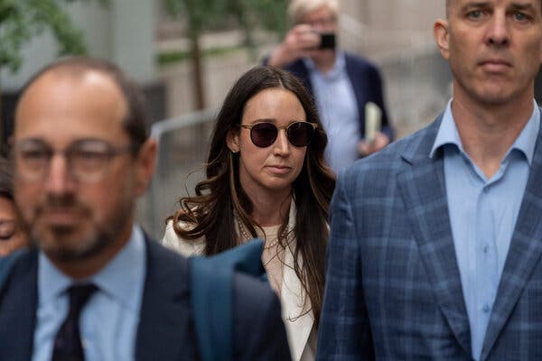 Charlie Javice with long, dark brown hair, wearing sunglasses and a white outfit, walking between two blurred men in suits.