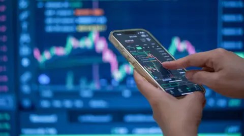 Getty Images A trader's hands using their phone to make a trade in front of a trading screen in soft focus