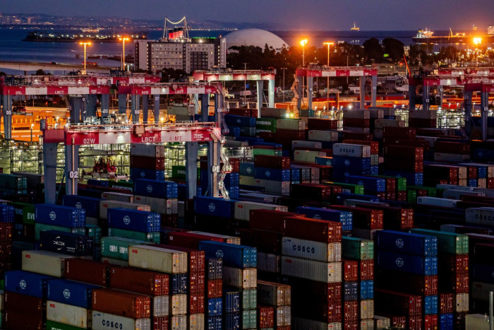 Shipping containers at the Port of Long Beach with large cranes visible, illuminated at dusk.