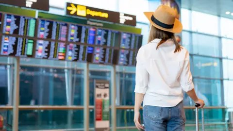 Getty Images Young woman at an airport wearing blue jeans, a white shirt and light brown panama hat stands with her back to the camera facing a flight departures board