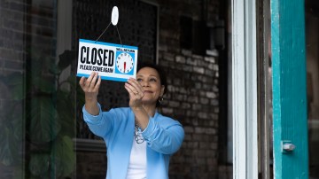 A woman holds a "CLOSED, Please come again" sign.