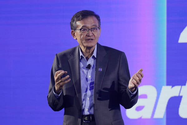 Lip-Bu Tan wears a dark suit jacket over a blue shirt, with a small microphone attached to his shirt. He is speaking and gesturing.