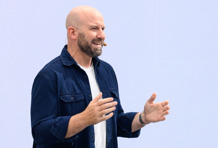 Andrew Bosworth, Meta CTO, is mid speech with his hands held aloft in front of him, as if emphasizing a point. He is standing in front of a light blue background and wearing a dark denim overshirt, which is open over a white undershirt. He is wearing a microphone on his face.