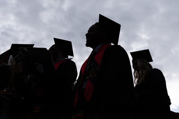College graduates, wearing gowns and mortarboards, are seen in silhouette against a cloudy sky. 
